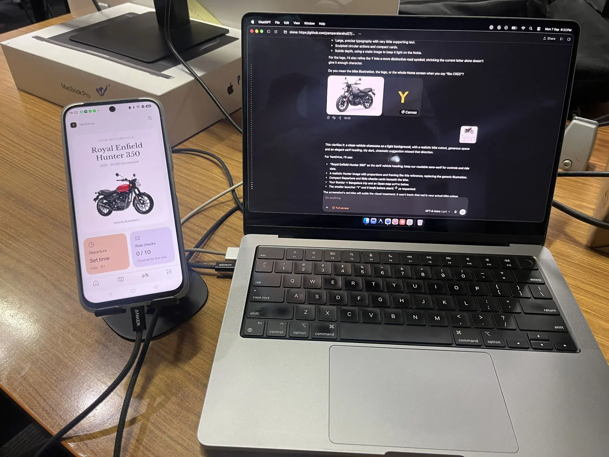 YamDrive running on an Android phone in a desk stand, showing the Royal Enfield Hunter 350 home screen with Departure and Ride checks cards, beside a MacBook Pro with the AI coding session that is building the app