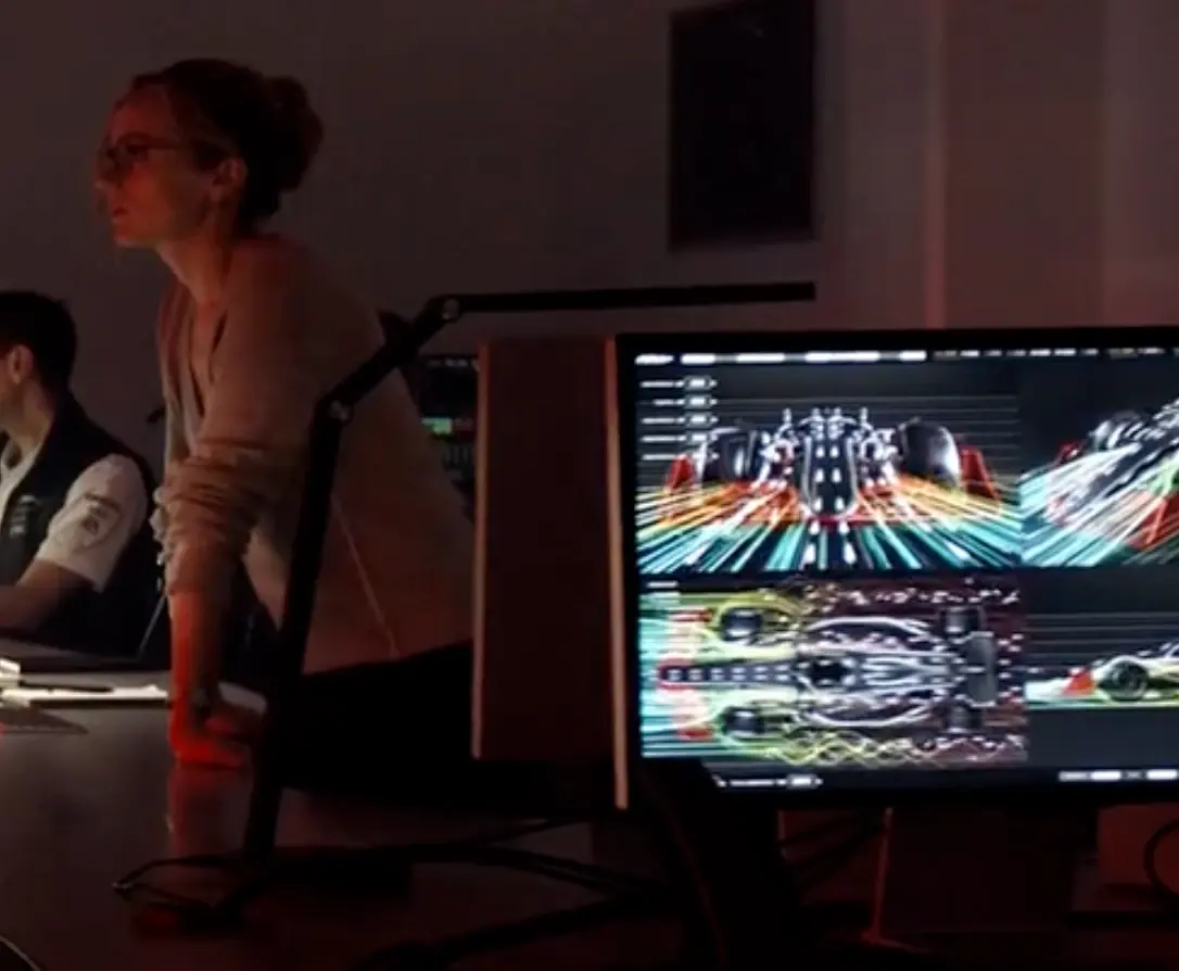 Still from F1 (2025): an engineer in a dark room leaning over a desk beside a monitor showing the car in four aerodynamic views, front, top and side, with airflow drawn as coloured streaks