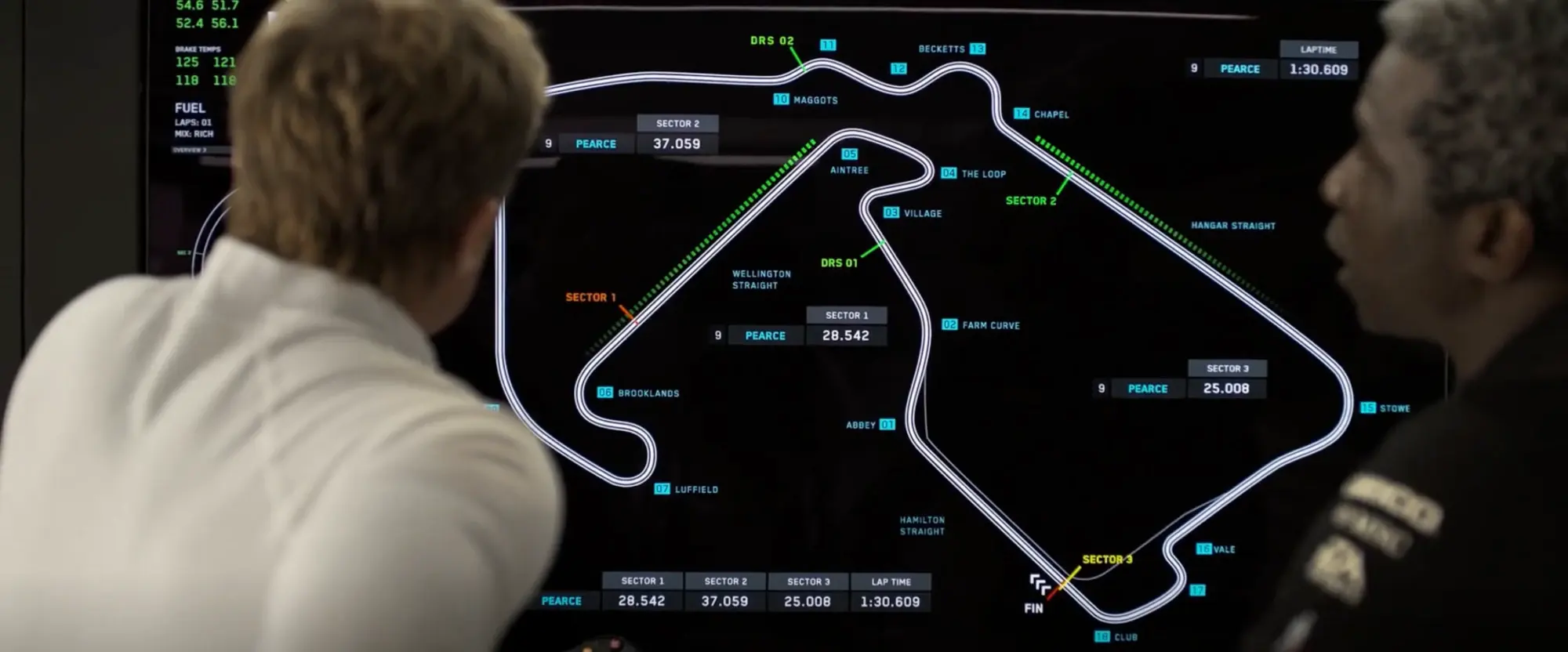 Still from F1 (2025): two people at a pit wall screen showing the whole Silverstone circuit with every corner numbered and named, DRS zones and sector lines marked, a time card beside each sector for Pearce, a lap time of 1:30.609 top right and the completed split table below