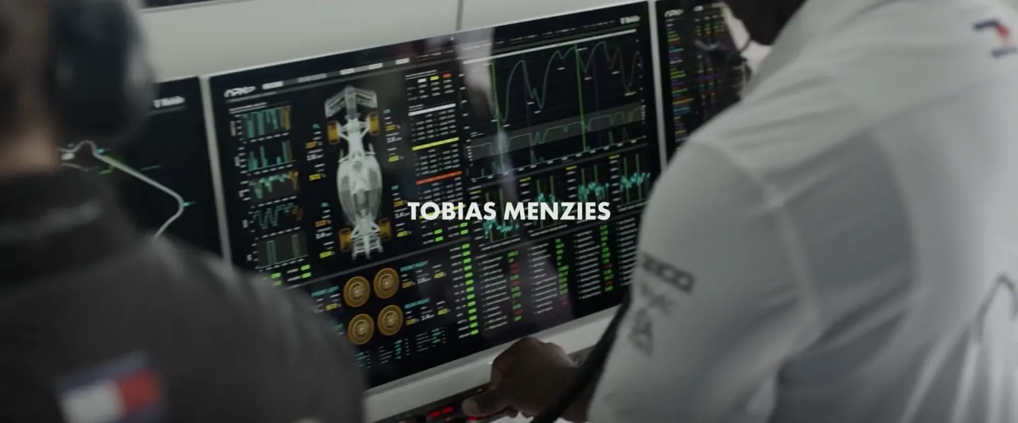 Still from F1 (2025): APXGP engineers at the pit wall in front of a telemetry screen with a top-down car diagram, four tyre readouts, stacked bar charts and live line traces