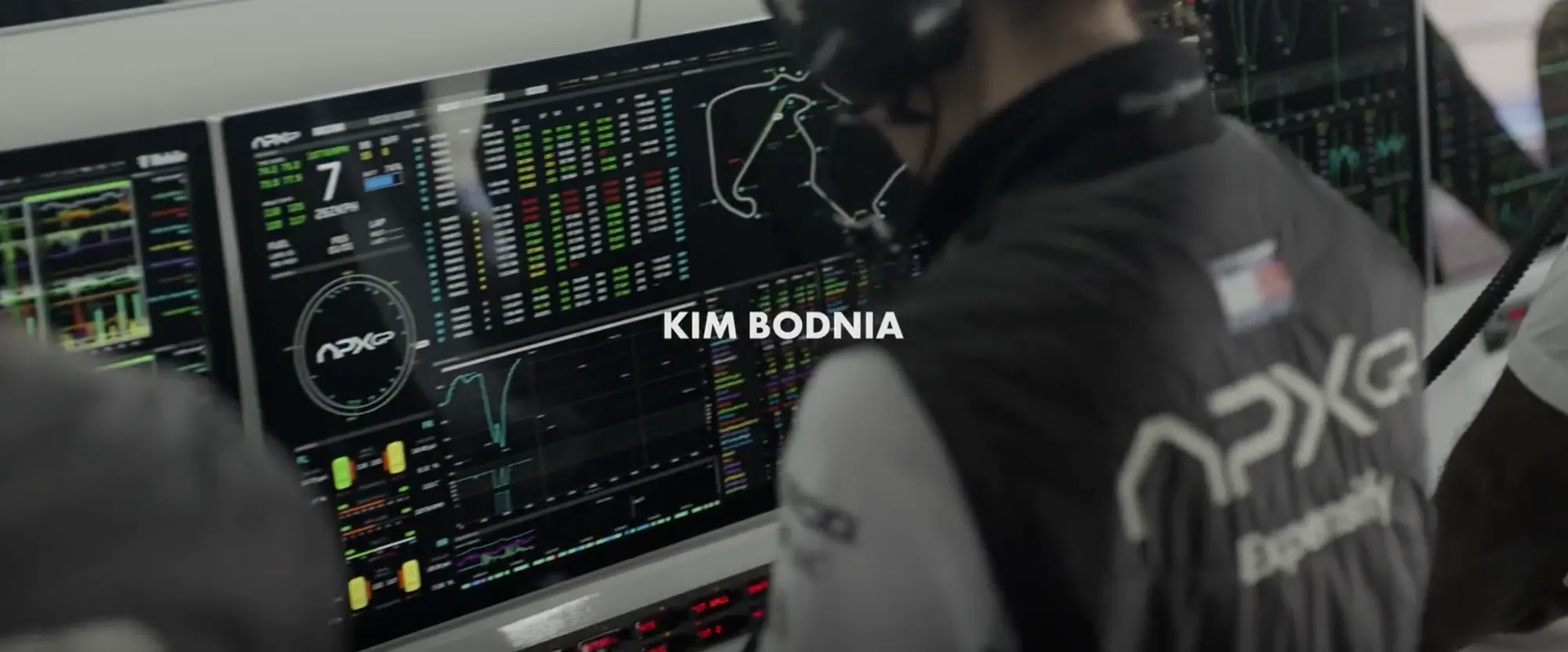Still from F1 (2025): an APXGP engineer at the pit wall in front of a screen with a large gear number and speed, a timing grid in green, yellow and red, a track map, tyre state pills and a lap trace