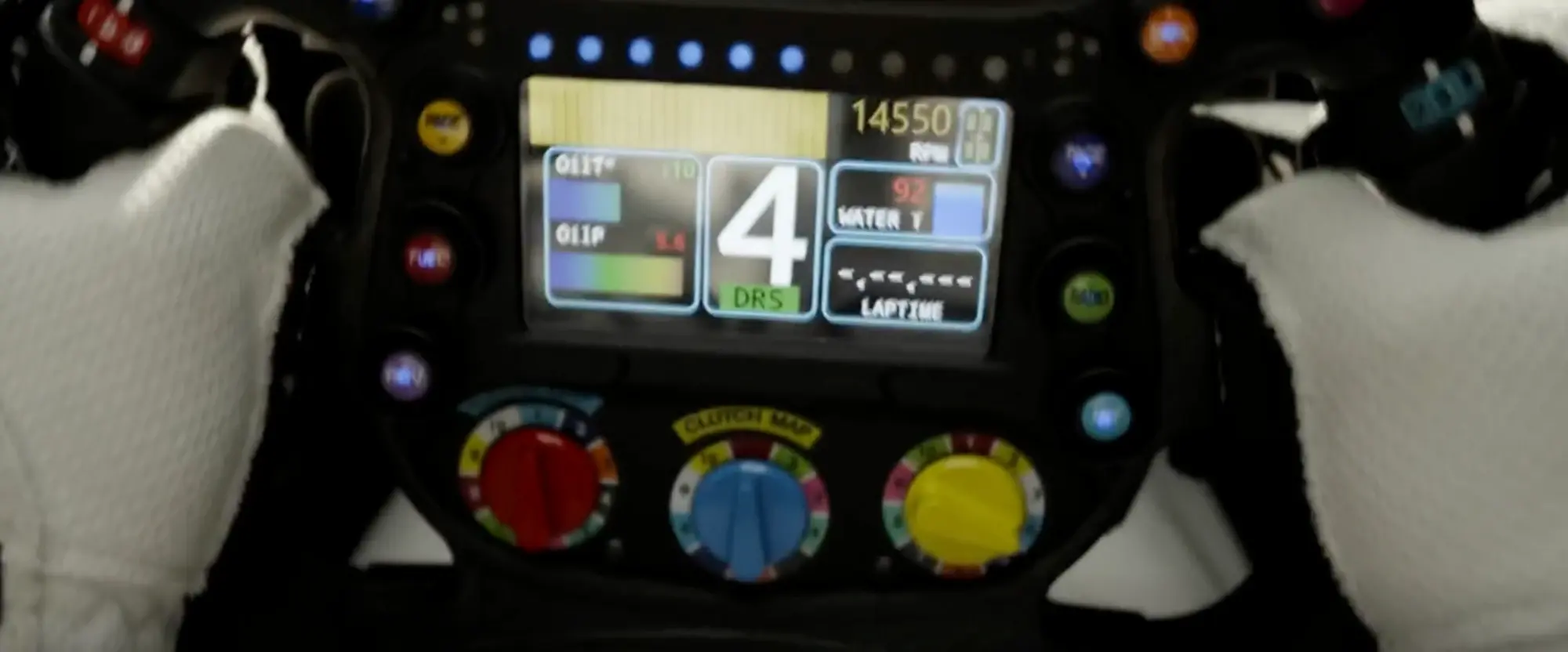 Still from F1 (2025): a driver's gloved hands on an F1 steering wheel whose screen shows gear 4 with a DRS badge, 14550 RPM, oil and water temperatures and lap time, above a row of blue shift lights and three colour-coded rotary dials