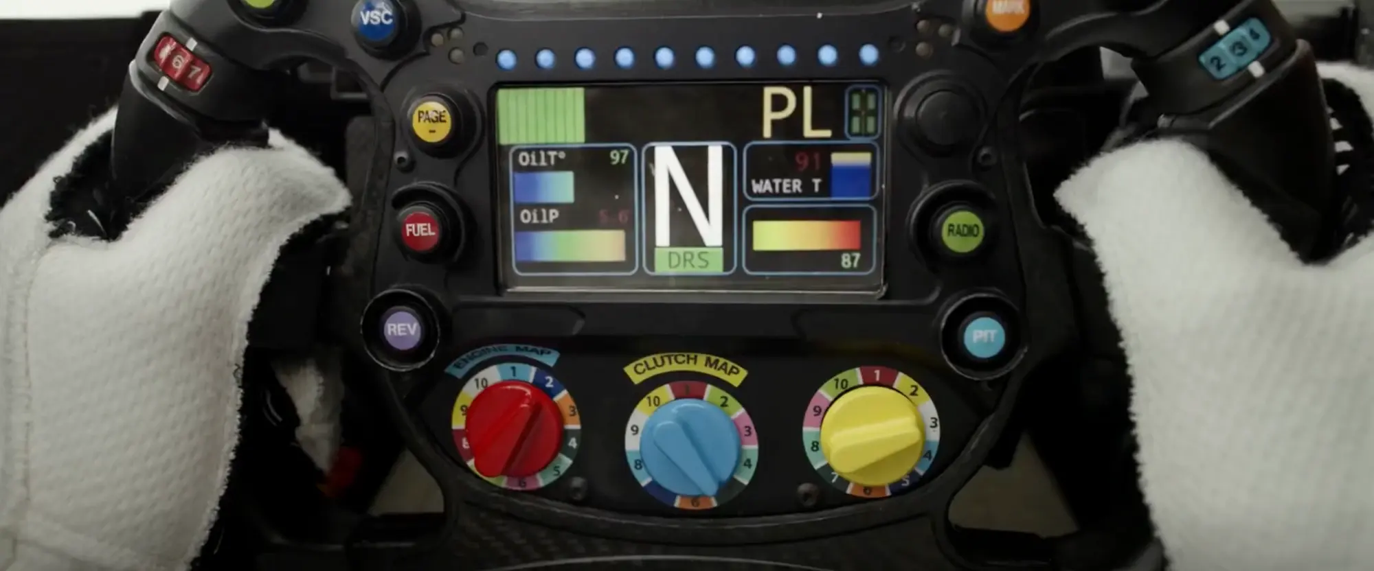 Still from F1 (2025): a sharp view of the steering wheel screen showing N for neutral with a DRS badge, PL for pit limiter, oil temperature and pressure as gradient bars, water temperature, and labelled buttons for Page, Fuel, Rev, Radio and Pit above three colour-coded rotary dials